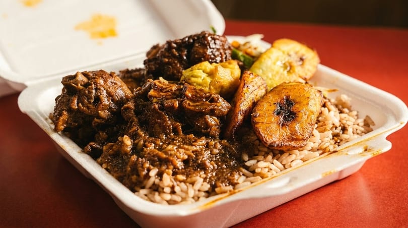 Oxtail stew with rice and peas, fried plantains, and dumplings in a takeout container.