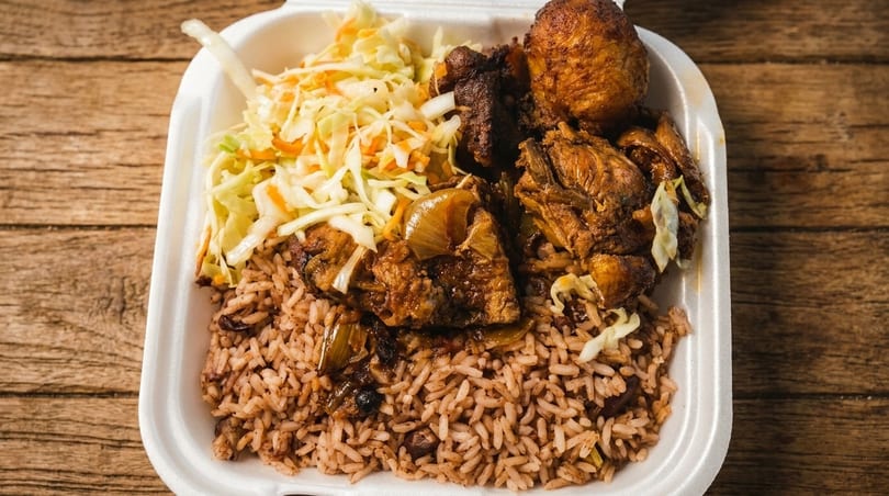 A takeout container filled with brown rice, stewed chicken, and coleslaw on a wooden table.